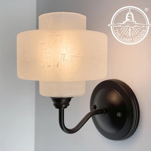 May include: A wall-mounted light fixture with a frosted glass shade and a dark bronze finish. The lamp has a circular base and a curved arm. The lamp is designed for indoor use and is suitable for a variety of spaces.