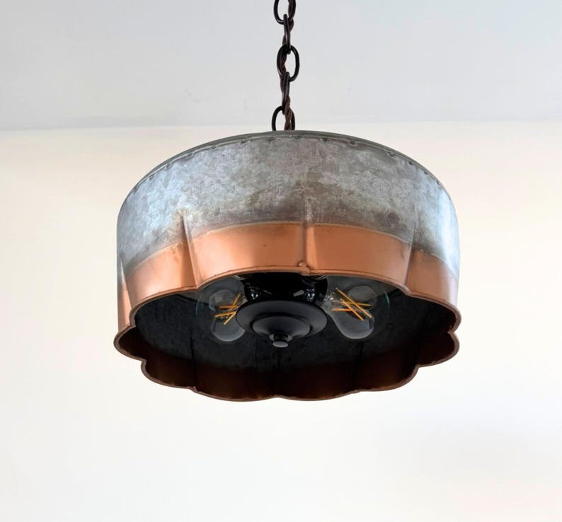 May include: A hanging pendant light with a galvanized metal shade and copper-colored band. The light fixture has a scalloped edge and three visible light bulbs. The fixture hangs from a dark chain.