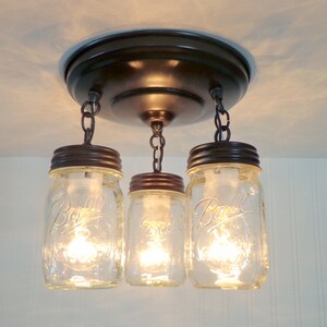 May include: A three-light fixture with clear glass mason jar shades. The jars are suspended from a bronze-colored ceiling mount by chains. Each jar has a bronze-colored lid and contains a lit bulb, creating a warm glow.