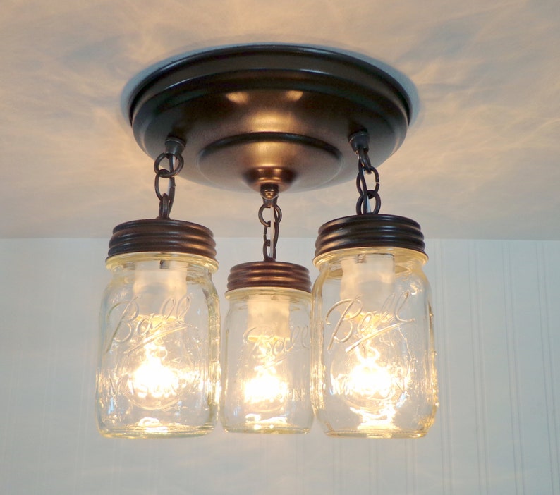 May include: A three-light fixture with clear glass mason jar shades. The jars are suspended from a bronze-colored ceiling mount by chains. Each jar has a bronze-colored lid and contains a lit bulb, creating a warm glow.