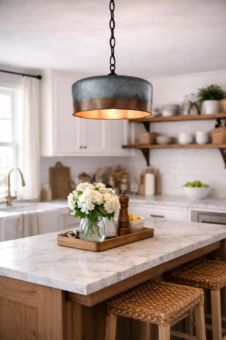 May include: A rustic kitchen pendant light with a galvanized metal shade and copper trim hangs above a marble countertop. A wooden tray holds a vase of white flowers, a pepper grinder, and lemons. Woven stools sit below.