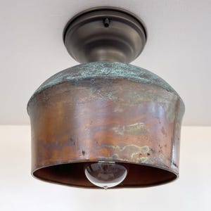 May include: A vintage-style ceiling light fixture with a copper-colored shade and a bronze-colored base. The shade has a rounded, bell-like shape with a clear lightbulb visible at the bottom. The copper has a patina with hints of green.