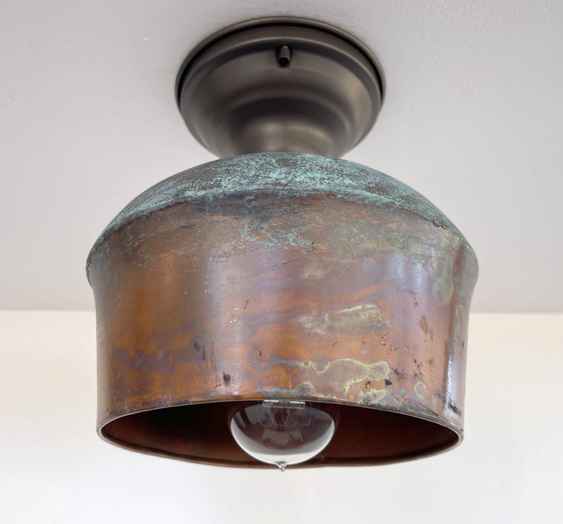 May include: A vintage-style ceiling light fixture with a copper-colored shade and a bronze-colored base. The shade has a rounded, bell-like shape with a clear lightbulb visible at the bottom. The copper has a patina with hints of green.