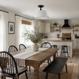 May include: A dining room featuring a large wooden table with a light-colored runner, surrounded by black spindle chairs and a black bench. A white ceramic vase with greenery sits on the table under a white drum pendant light with black trim.