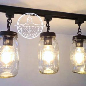 May include: Four glass mason jars with light bulbs inside are hanging from a black track lighting system. The jars are clear glass with a textured surface and have black metal lids. The track lighting system is mounted to the ceiling.