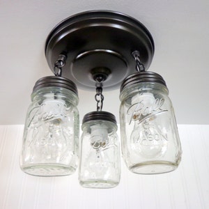 May include: A rustic ceiling light fixture featuring three clear glass mason jars with the "Ball Mason" logo, suspended from a dark bronze-colored metal base. The jars are lit with bulbs, creating a warm, inviting glow.