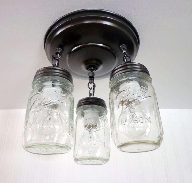 May include: A rustic ceiling light fixture featuring three clear glass mason jars with the "Ball Mason" logo, suspended from a dark bronze-colored metal base. The jars are lit with bulbs, creating a warm, inviting glow.