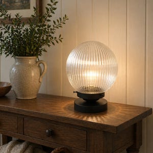 Peut inclure: Lampe sphérique en verre transparent et strié, posée sur une table en bois. La base est noire. La lumière chaude de la lampe se diffuse. Un vase avec des plantes vertes est visible en arrière-plan, évoquant une ambiance décorative.
