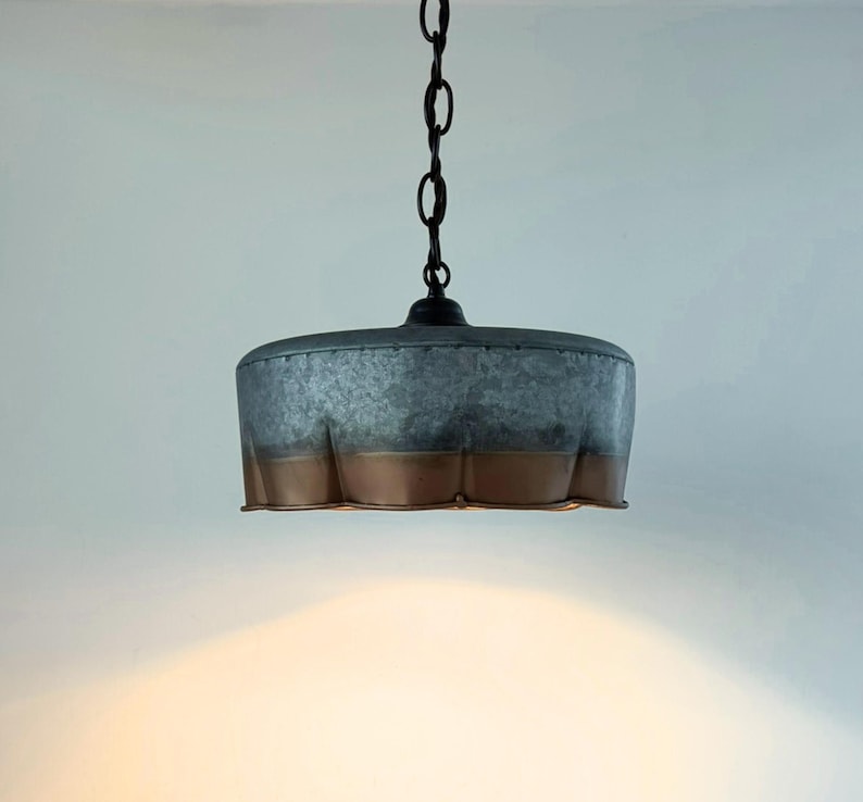 May include: A hanging pendant light with a galvanized metal shade and a copper-colored bottom edge. The light fixture is suspended from a black chain and is suitable for indoor lighting. The shade has a unique, textured appearance.