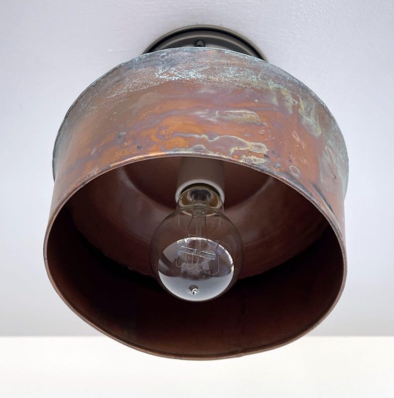 May include: A vintage-style ceiling light fixture with a copper-colored shade. The shade has a cylindrical shape with a distressed finish, revealing hints of green and brown. A clear light bulb is visible inside the shade, and the fixture is mounted on a white ceiling.