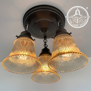 May include: A three-light flush mount ceiling fixture with a dark bronze finish and clear, ribbed glass bell-shaped shades. Each shade features a beaded edge and houses an exposed filament bulb. The logo for The Lamp Goods is visible.