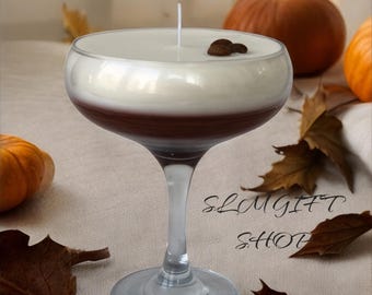 May include: A white and brown layered candle in a martini glass with coffee beans on top. The candle is on a beige surface with brown leaves and the text "SLUMGIFT SHOP".