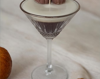 May include: A white candle in a martini glass with two brown pumpkin-shaped candle toppers. The glass is on a white fabric surface with small orange pumpkins around it.