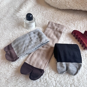 May include: Three pairs of split-toe socks in grey, tan, and black with brown toe sections. A small perfume bottle with the text "BYREDO MOJAVE GHOST" and a burgundy hair clip are also visible on a white, textured surface.