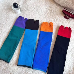 Op de afbeelding: Vier paar kleurrijke tabi-sokken met een split-teenontwerp liggen op een witte, zachte ondergrond. Elk paar heeft een colorblock-patroon in de kleuren teal, paars, zwart, blauw, oranje en rood.