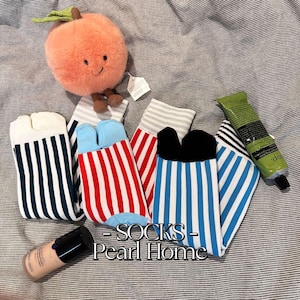 May include: Four pairs of striped socks with split toes. The socks are in blue, red, white, and black. The socks are arranged on a light gray fabric background. The text "SOCKS - Pearl Home" is printed on the fabric.