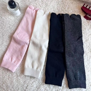 Peut inclure: Quatre paires de chaussettes tabi roses, blanches, bleu marine et grises. Les chaussettes sont côtelées et ont un design à bout fendu. Un flacon de parfum et une pince à cheveux rouge sont en arrière-plan.