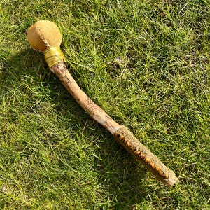 May include: A handcrafted wooden mace with a tan leather head and a decorative handle. The handle is wrapped with a gold-colored band and adorned with small, round, brown beads. The mace is lying on a bed of green grass.
