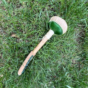 May include: A maraca, a percussion instrument, lies on green grass. It has a light brown wooden handle, a tan and green head, and blue beads along the handle. The maraca is handmade.