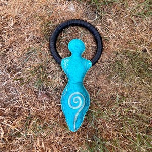 May include: A turquoise fabric figure with a rounded head and a wide, curved body, featuring a central spiral design and white line accents. The figure is attached to a circular black ring that frames the upper portion of the object.