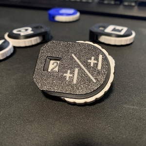 May include: A collection of black and white game tokens with various symbols and numbers. The central token displays the number 2, along with +1 symbols. The tokens have a textured surface and a white rim, suggesting they are 3D printed.