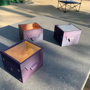 Puede incluir: Tres pequeñas cajas cuadradas con detalles morados y dorados. Cada caja tiene una parte superior diferente: una es de color cobre, otra transparente y otra dorada. Las cajas están sobre una superficie gris.