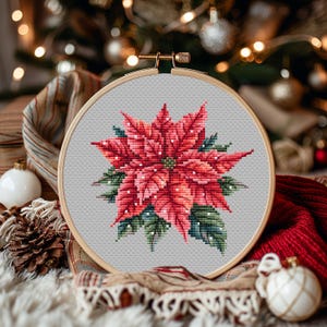 Pode incluir: Um bordado em ponto cruz de uma flor de poinsétia num aro de madeira. A flor tem pétalas vermelhas e rosa com folhas verdes e detalhes brancos. O fundo é um tecido cinza claro.
