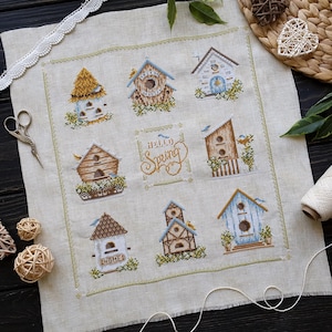 May include: A cross-stitch sampler featuring nine birdhouses in various designs and colours, with the text "Hello Spring" in the centre. The birdhouses are surrounded by greenery and small birds, all on a beige linen fabric.