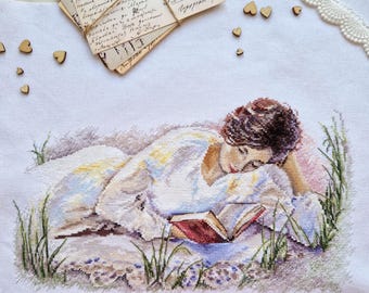 May include: A cross-stitch embroidery of a woman lying in a field of grass, reading a book. The woman is wearing a white dress and has brown hair. The book is red and has a gold design on the cover.