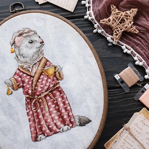 May include: Cross-stitch embroidery of a ferret wearing a red dressing gown and holding a cup, framed in a wooden hoop.