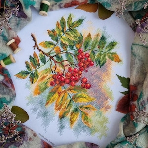 Könnte beinhalten: Kreuzstich-Stickerei eines Zweigs mit Ebereschenbeeren mit grünen und gelben Blättern. Die Beeren sind rot und der Hintergrund ist hellblau.
