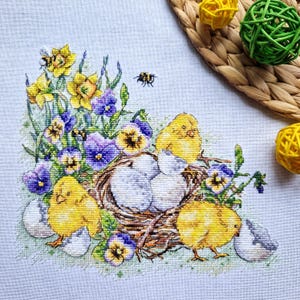 May include: Cross-stitch embroidery featuring a spring theme. A nest with speckled eggs is surrounded by yellow chicks, purple pansies, and yellow daffodils. Two bees are also present. The design is on a light blue fabric.