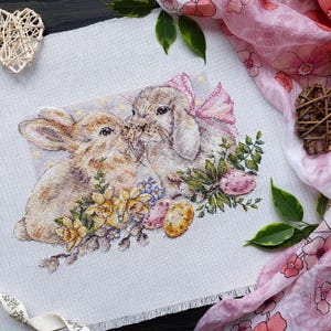 Puede incluir: Bordado de punto de cruz de dos conejos besándose, rodeados de flores y huevos de Pascua. Un conejo tiene un lazo rosa. La obra de arte está sobre un fondo de tela blanca, con una bufanda floral rosa y un corazón decorativo.