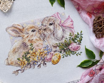 May include: Cross-stitch embroidery of two bunnies kissing, surrounded by flowers and Easter eggs. One bunny has a pink bow. The artwork is on a white fabric background, with a pink floral scarf and decorative heart.