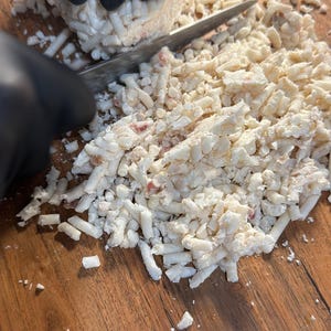 Op de afbeelding: Een hand met een handschoen gebruikt een mes om een ​​blok wit, kruimelig voedsel in kleine stukjes te hakken op een houten snijplank.