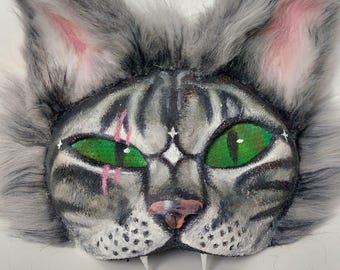 May include: A handmade cat mask with a gray and white striped face, bright green eyes, and pink accents. The mask has fluffy gray fur around the edges and pointed ears. The cat's face has a pink nose and white fangs.
