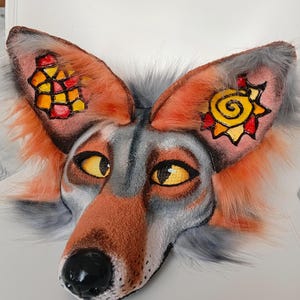 Può includere: Una maschera da animale simile a una volpe con pelliccia sintetica grigia e arancione, occhi gialli e naso nero. L'interno delle orecchie presenta disegni dipinti a mano: un motivo a griglia geometrica a sinistra e un sole a spirale a destra.
