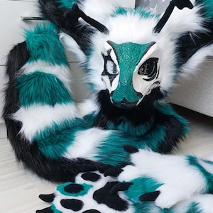 May include: A creature costume set featuring a mask with a green and white textured face, black and white eyes, and black antennae. The set includes long, striped teal, white, and black faux fur and matching paw gloves with black paw pads.