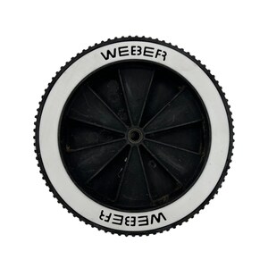 May include: A black and white wheel with the word "WEBER" printed in black on the white rim. The wheel has a black centre with spokes radiating outwards. The outer edge of the wheel has a ridged texture.