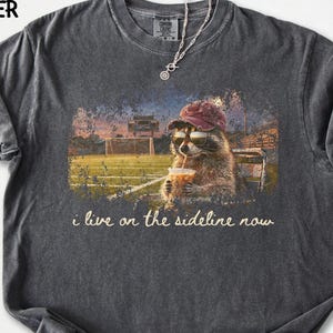 May include: Dark grey t-shirt featuring a raccoon wearing sunglasses and a cap, drinking from a cup with a straw. The graphic includes a football field scene and the text "i live on the sideline now". The shirt has a crew neck and short sleeves.