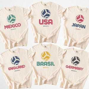 May include: Six cream-coloured t-shirts with soccer ball graphics and country names: Mexico, USA, Japan, England, Brazil, and Germany. Each shirt has a distressed print of a soccer ball and the text "WORLDCUP 24".
