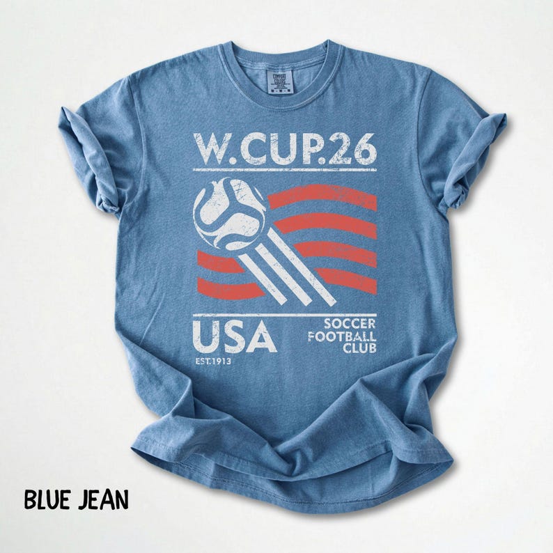 Flat lay of a blue jean color Comfort Colors t-shirt showcasing a vintage white and red USA Soccer Football Club 2026 design with a soccer ball.