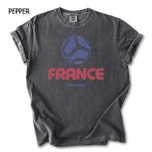 May include: A dark grey t-shirt featuring a blue football graphic and the word "FRANCE" in red. Below the word "FRANCE" is the text "WORLDCUP 26". The word "PEPPER" is in the top left corner of the t-shirt.