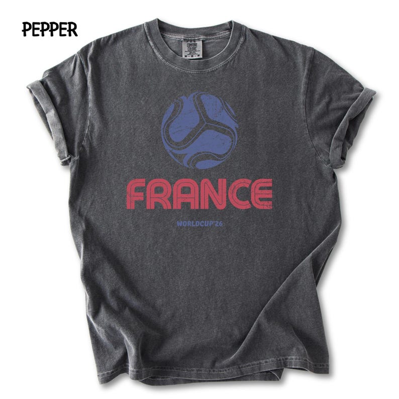 May include: A dark grey t-shirt featuring a blue football graphic and the word "FRANCE" in red. Below the word "FRANCE" is the text "WORLDCUP 26". The word "PEPPER" is in the top left corner of the t-shirt.