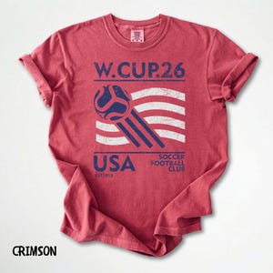 Flat lay view of a crimson red garment dyed t-shirt with a navy blue and white retro USA Soccer 26 logo and athletic stripes.