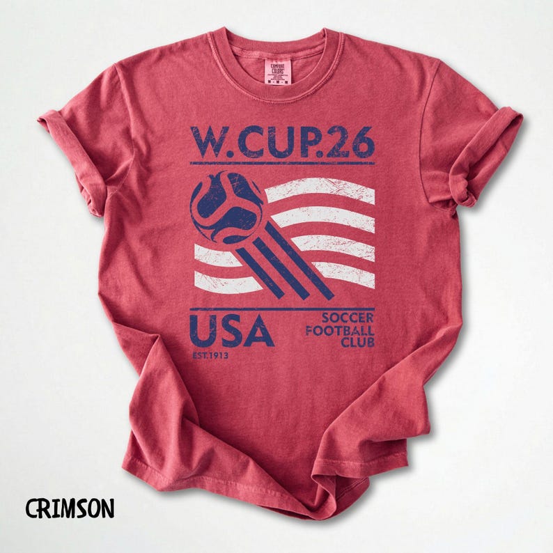 Flat lay view of a crimson red garment dyed t-shirt with a navy blue and white retro USA Soccer 26 logo and athletic stripes.