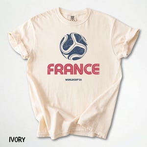 May include: An ivory-coloured t-shirt with a distressed navy and white football graphic above the word "FRANCE" in red, with "WORLDCUP'26" below. The shirt has a crew neck and rolled sleeves.