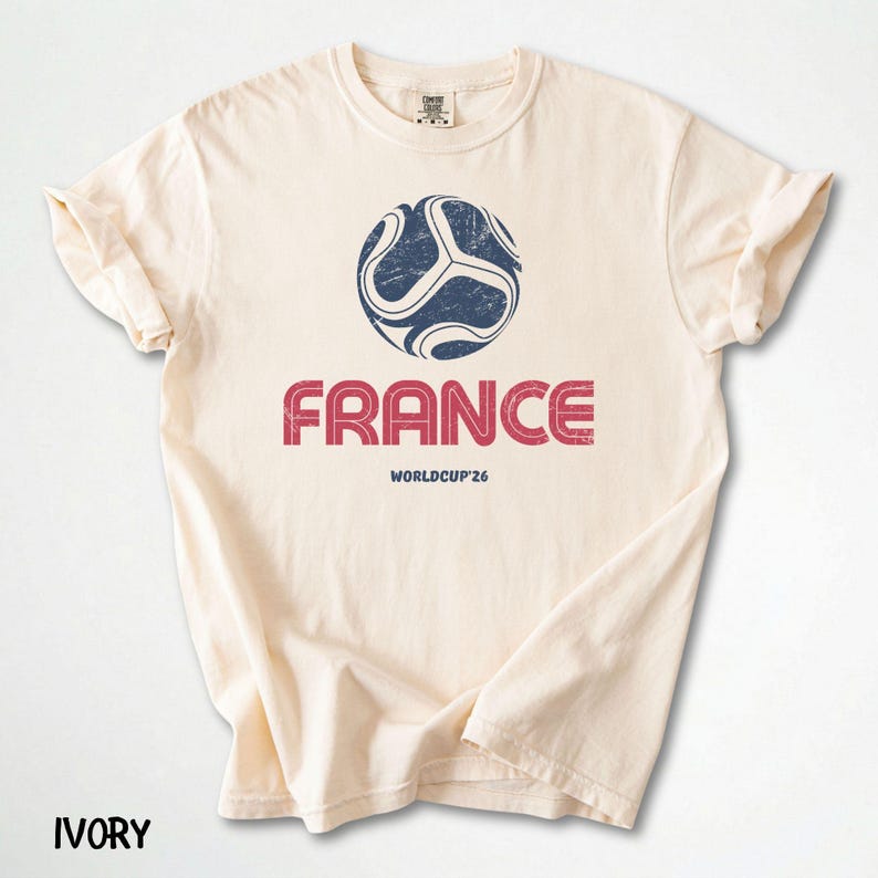 May include: An ivory-coloured t-shirt with a distressed navy and white football graphic above the word "FRANCE" in red, with "WORLDCUP'26" below. The shirt has a crew neck and rolled sleeves.