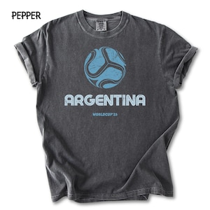 May include: Charcoal grey t-shirt with a faded design. It features a light blue football graphic above the word "ARGENTINA" and "WORLDCUP'26". The shirt has rolled sleeves and the word "PEPPER" is printed at the top left.