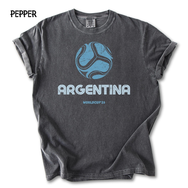 May include: Charcoal grey t-shirt with a faded design. It features a light blue football graphic above the word "ARGENTINA" and "WORLDCUP'26". The shirt has rolled sleeves and the word "PEPPER" is printed at the top left.